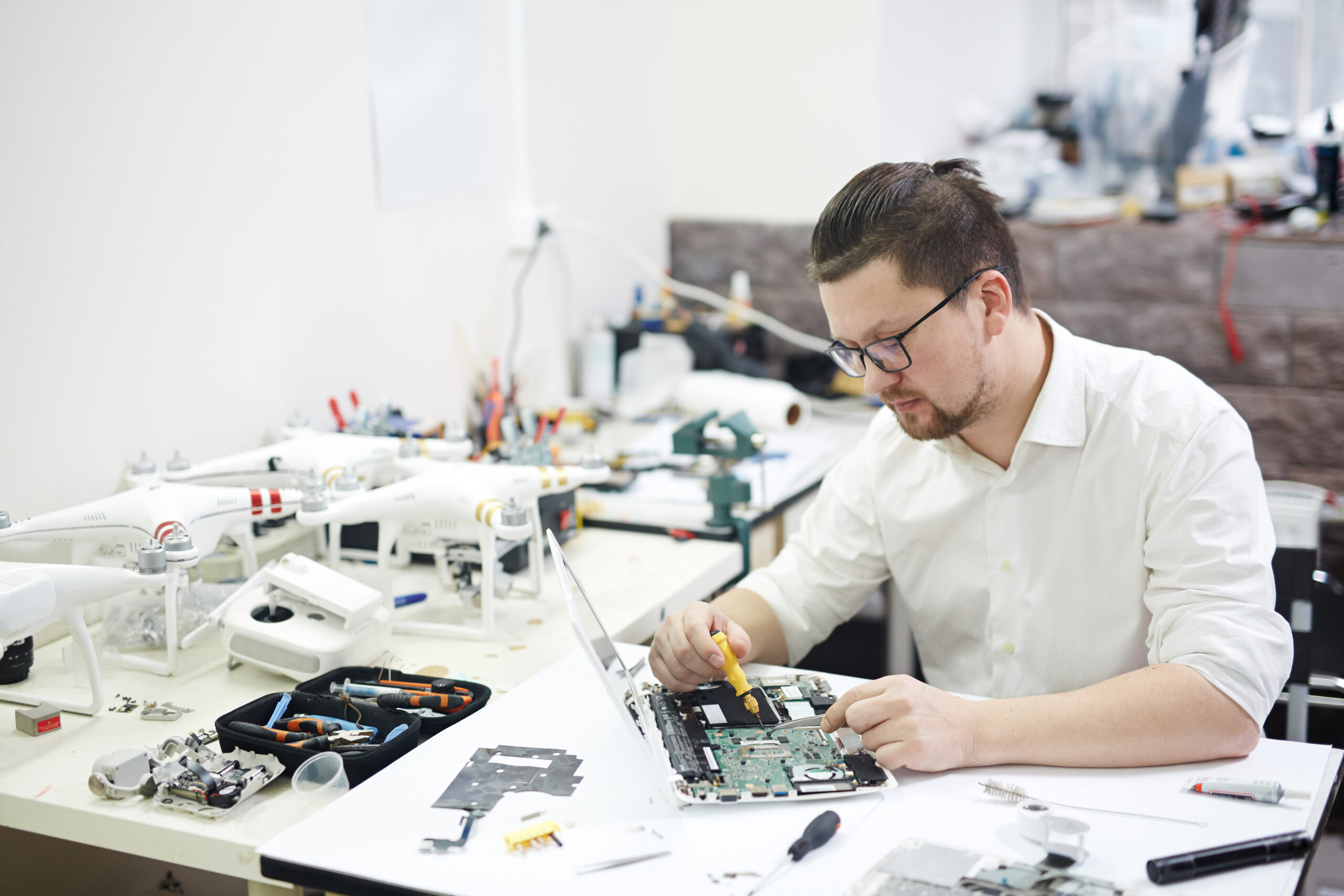 Portrait of modern man wearing glasses busy working with electronics in workshop: disassembling laptop and drones with different tools and electronic devices on table